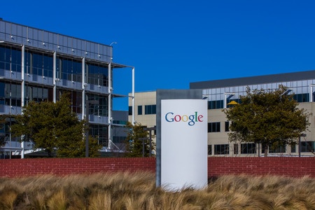This is an exterior view of Google's corporate headquarters in Mountainview, California. The building looks cool and futuristic and there is a large white sign that says Google in front of the building.