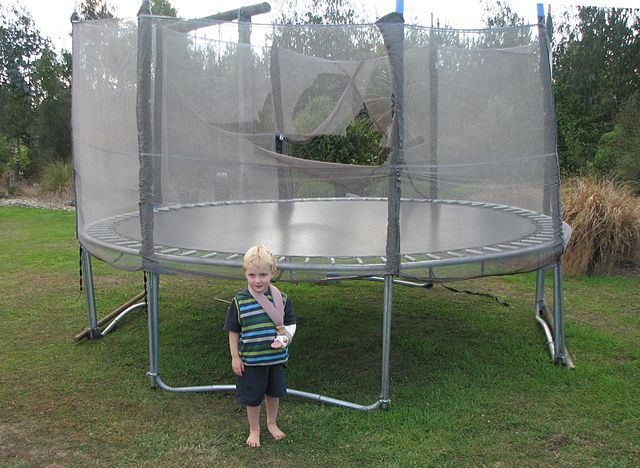 This is a young boy with his arm in a sling. He is standing next to a trampoline with safety nets. One of the nets has a large hole in it.