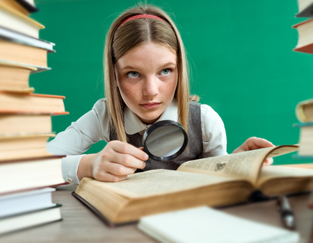 This is an image of a young girl holding a magnifying glass to an open book. She appears to be deep in thought and is surrounded by a pile of books.