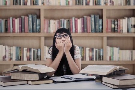 This is an image of a young woman in a library. She is sitting at a desk with a pile of books and looking toward the ceiling. She appears to be in deep thought.